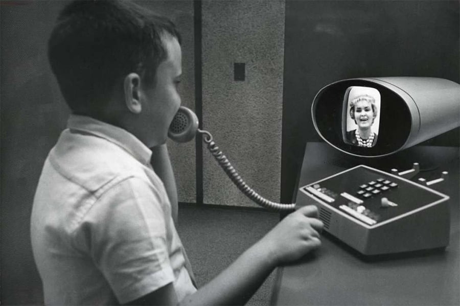 A boy uses a vintage video phone, holding a handset while a woman’s face appears on the screen. The device has buttons and a sleek, retro design.