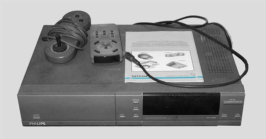 A black and white photo of a vintage Philips CD-i player, with a wired joystick, remote control, cord, and instruction manual placed on top.