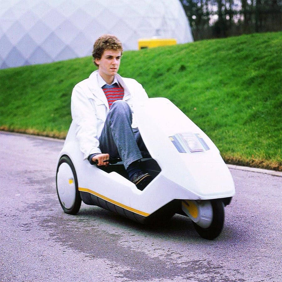 A person drives a small, white, futuristic three-wheeled electric vehicle on a paved road beside green grass and a dome-shaped building.