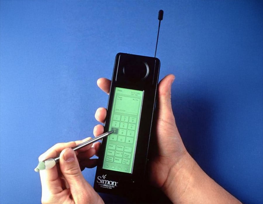 A person uses a stylus on the touchscreen of an IBM Simon, an early smartphone with a large antenna, against a blue background.