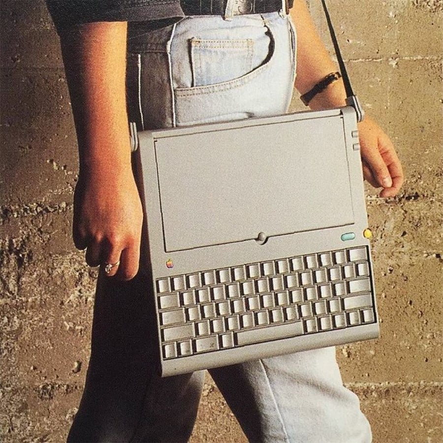 A person wearing light blue jeans carries a vintage laptop by its screen, using it like a briefcase, against a textured concrete wall.