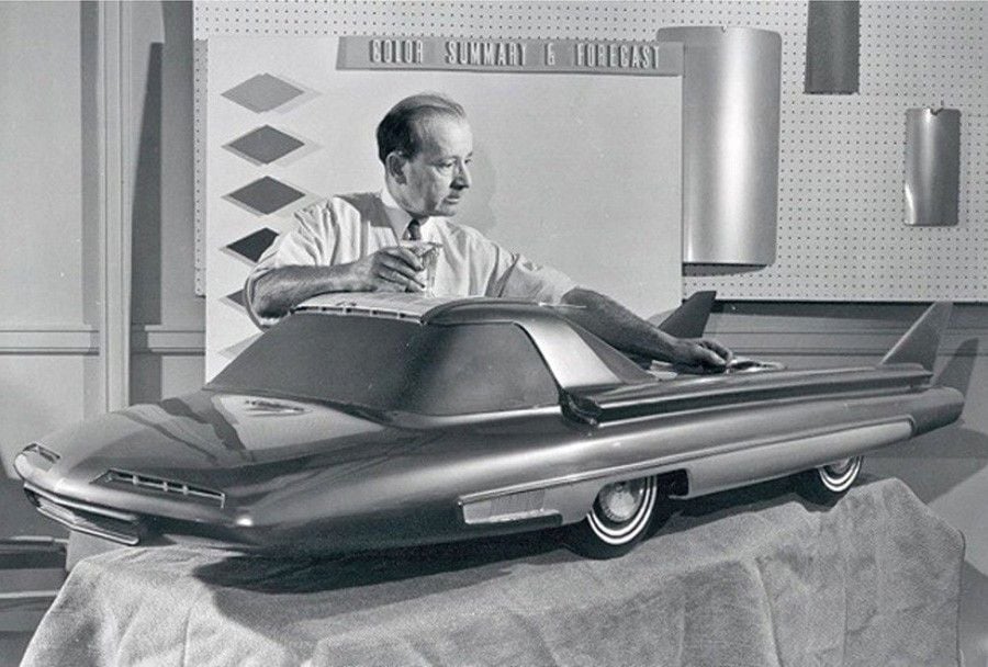 A man adjusts a futuristic car model on a table, with color samples and charts displayed on the wall behind him. The scene is in black and white.