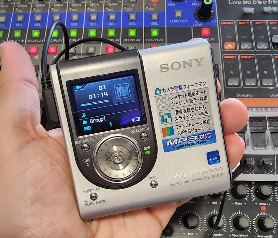 A hand holds a silver Sony Hi-MD Walkman digital music player, displaying a song on its screen. An audio mixing console with colorful buttons and sliders is in the background.