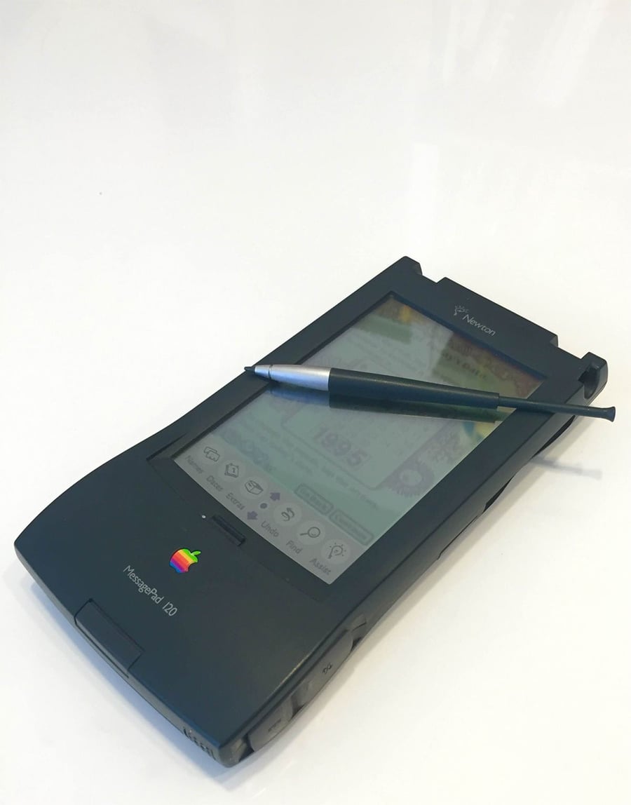 An Apple Newton MessagePad 120 PDA with a stylus and pen resting on its screen, displayed on a white surface.