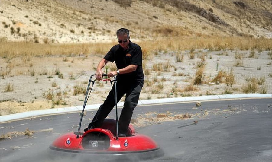A man in sunglasses rides a small red hovercraft over a dry, dusty surface, with barren hills and sparse grass in the background.