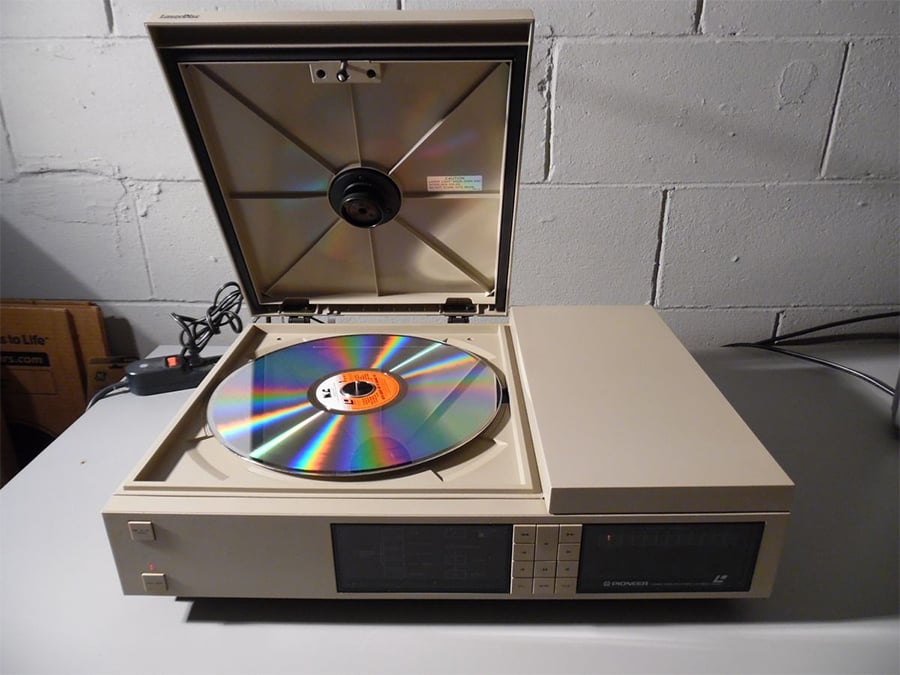 A vintage LaserDisc player with the lid open and a reflective disc inside sits on a gray table against a cinder block wall.