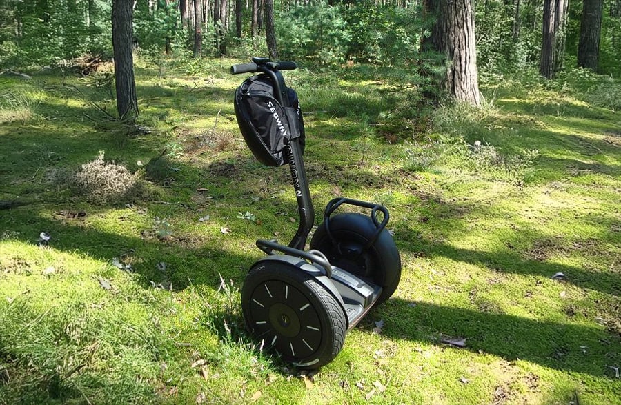 A black Segway with a handlebar bag stands on a sunlit, mossy forest floor surrounded by trees.