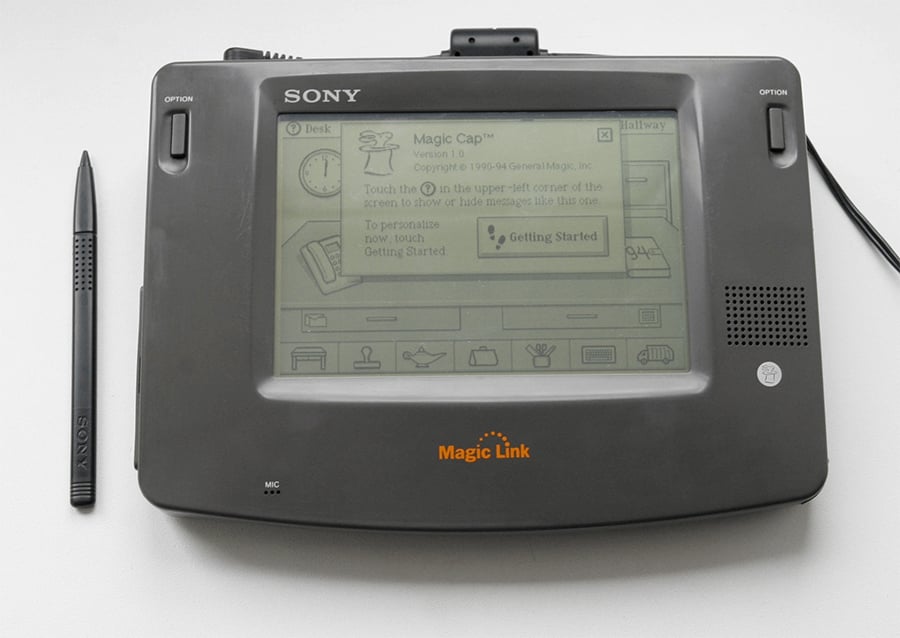 A black Sony Magic Link personal digital assistant with a stylus on the left. The screen displays a graphical user interface with icons and the text “Magic Cap” and “Getting Started.”