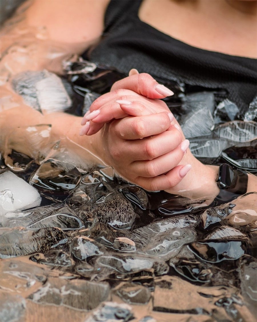 A person with clasped hands rests their arms in a tub filled with ice cubes, suggesting they are taking an ice bath. The person is wearing a black top, and only their hands and part of the arms are visible.