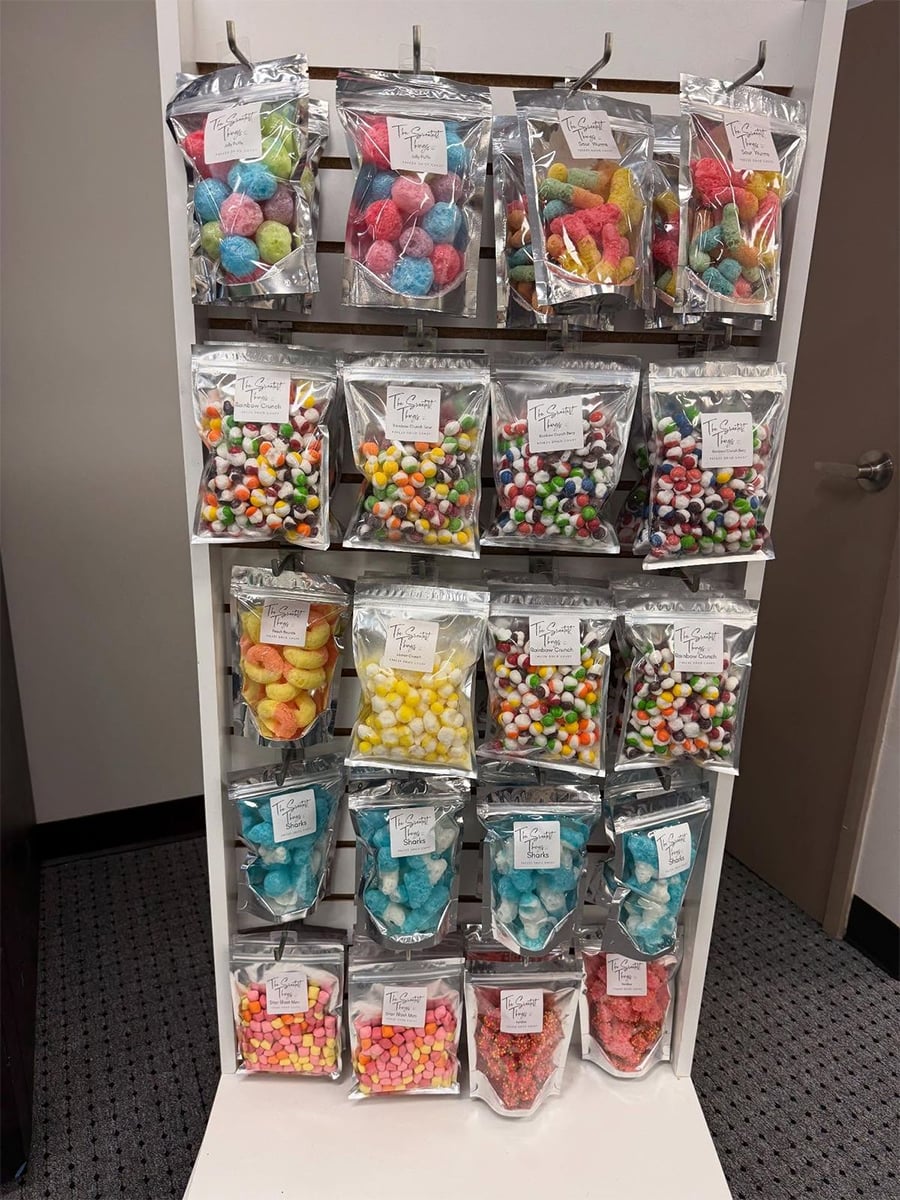 Display rack filled with various colorful packaged candies in clear resealable bags, arranged neatly in rows on metal hooks against a white background, with gray carpeted floor and a door in the background.