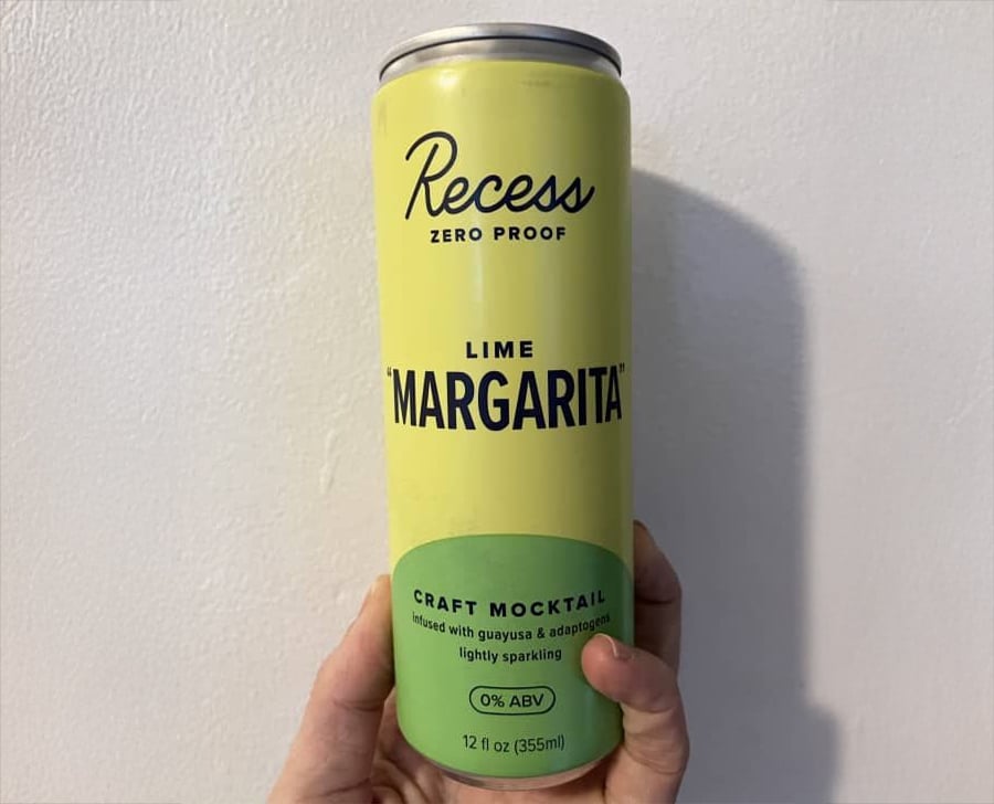 A hand holds a 12 oz can of Recess Zero Proof Lime Margarita craft mocktail with a yellow and green design against a plain wall background.