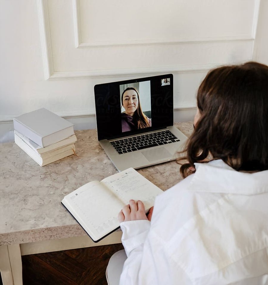 A person in a white shirt sits at a desk with books, taking notes in a notebook while having a video call on a laptop with another person.