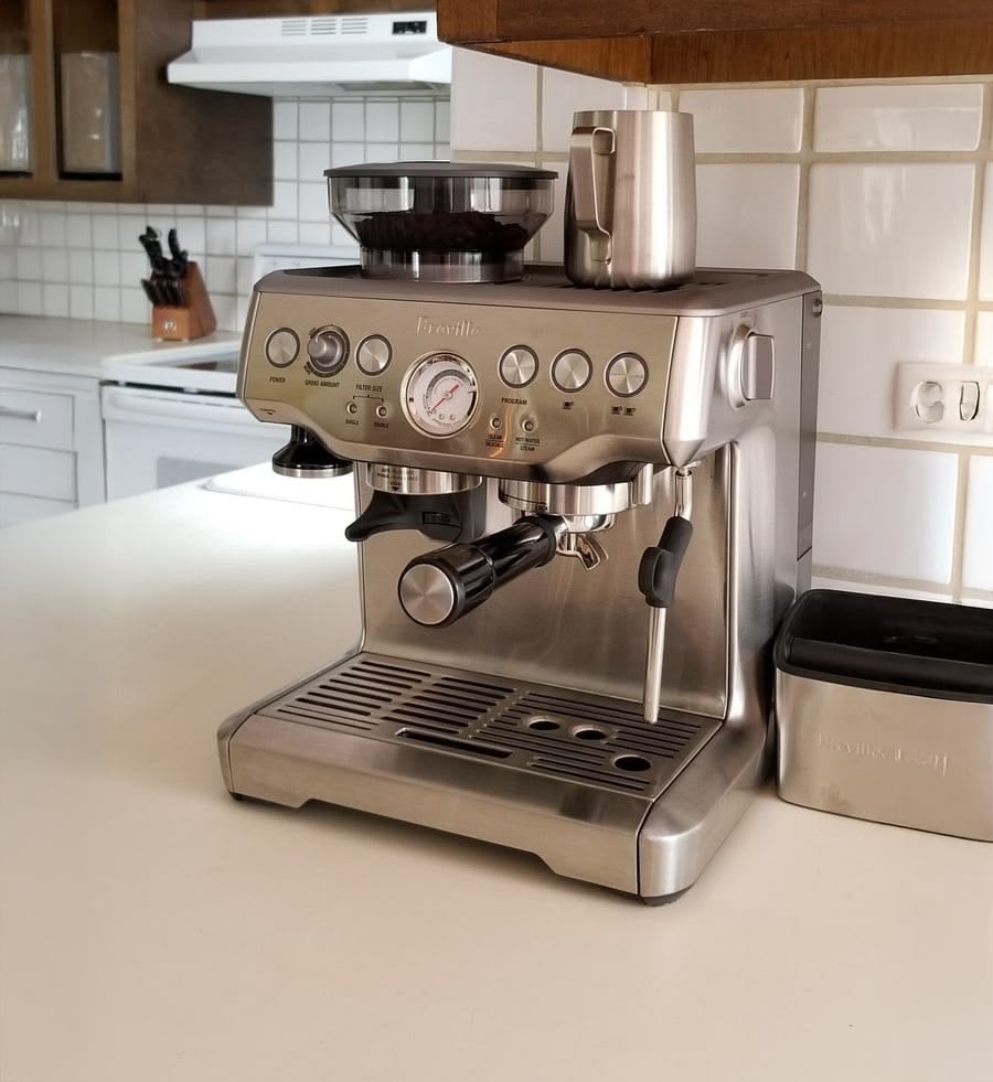 A silver Breville espresso machine with a milk frothing pitcher on top sits on a kitchen counter. White tile backsplash and drawers are in the background.