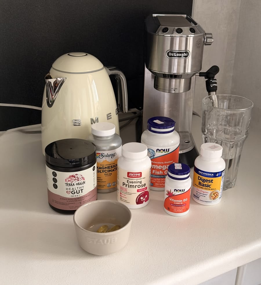 A kitchen counter with a kettle, espresso machine, glass, and assorted supplement bottles. A small bowl with capsules sits near the supplements.