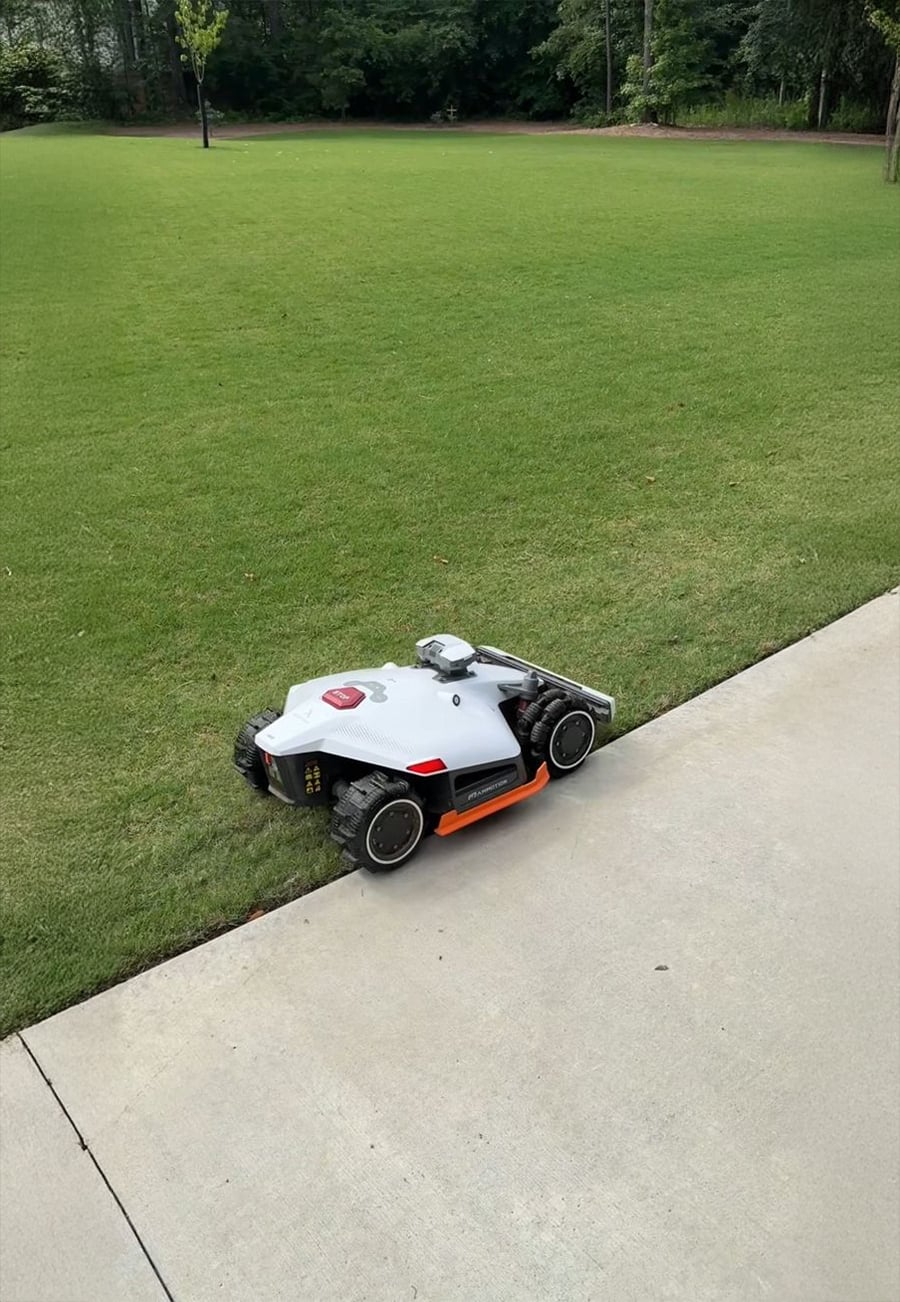 A robotic lawn mower sits on the edge of a concrete path and a grassy lawn, with trees and more grass in the background.