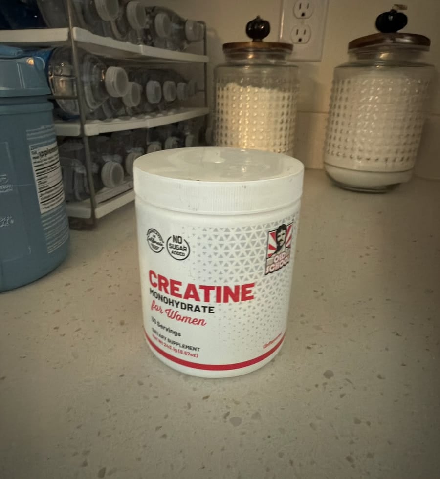 A white container of creatine monohydrate for women sits on a kitchen counter, with water bottles and two patterned jars with lids in the background.
