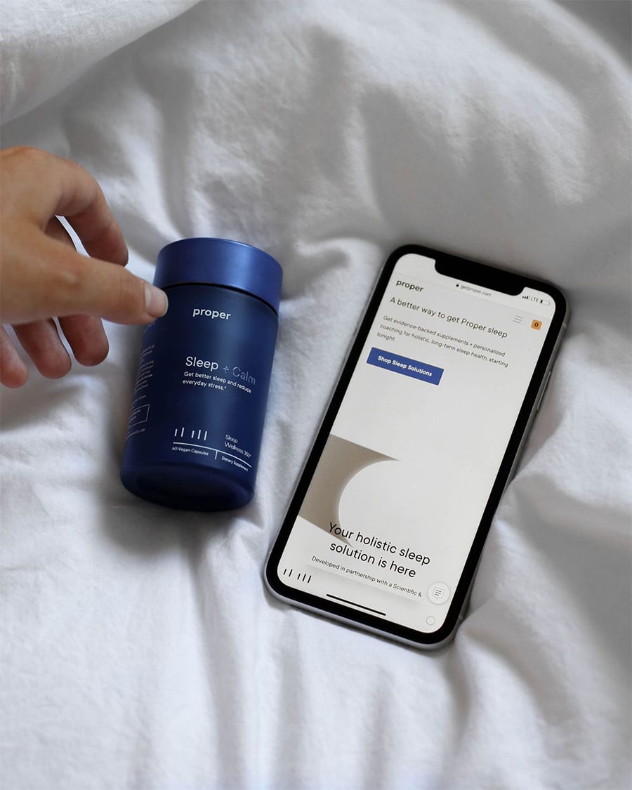 A hand reaches for a blue bottle labeled "Sleep + Calm" on a white bed, next to a smartphone displaying a sleep solution website.