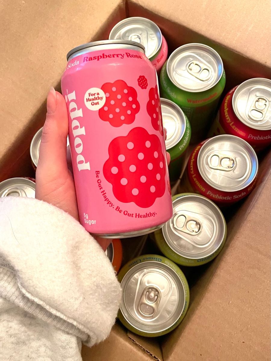 A hand holds a pink can of Poppi Raspberry Rose soda above a box filled with assorted cans of Poppi prebiotic soda in various colors.