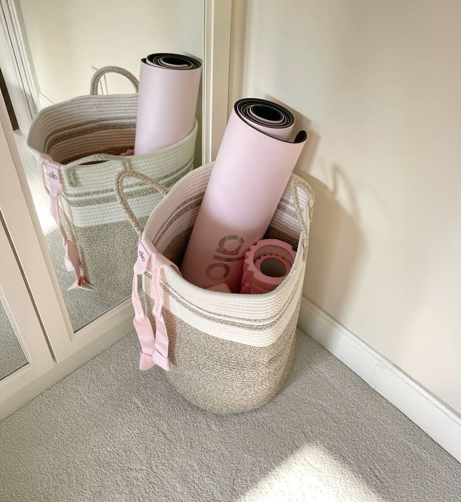 A woven basket holds a pink yoga mat and a pink foam roller, placed on a light carpet next to a mirrored closet and cream-colored wall.