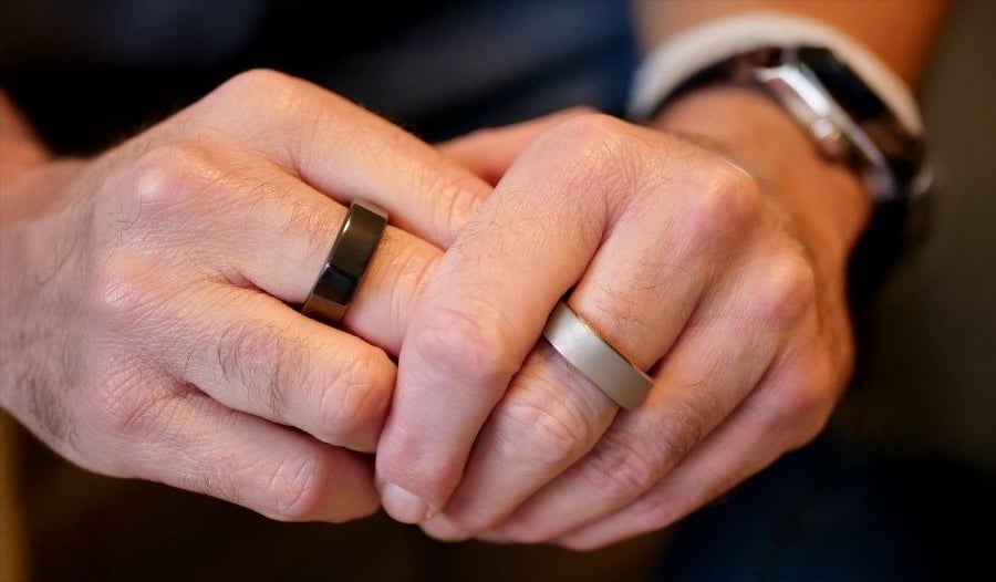 Two hands with interlaced fingers, each wearing a different smart ring—one black and one silver. A white watch is visible on the wrist in the background.