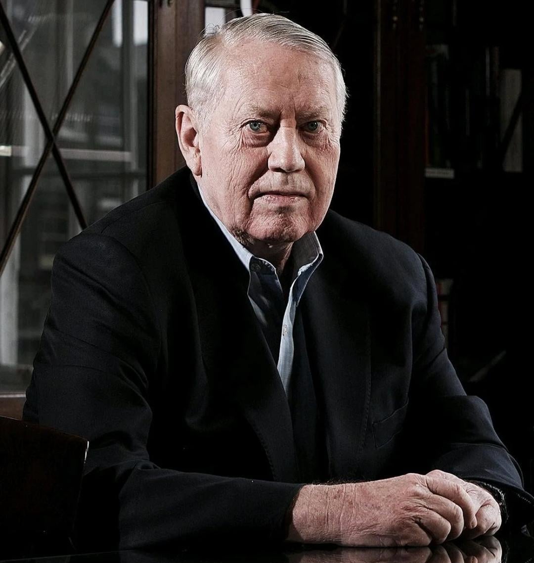 An older man with short gray hair wearing a dark suit sits at a table with his hands clasped, looking seriously at the camera. A bookshelf with glass doors is visible in the background.