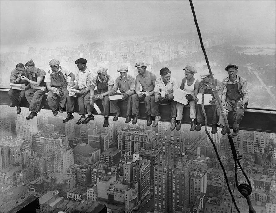 Eleven construction workers sit on a steel beam high above a city, eating lunch and chatting, with skyscrapers and streets visible far below.
