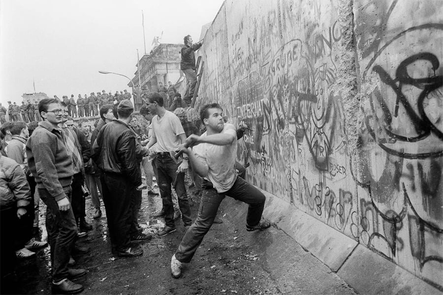 A man swings a hammer at the Berlin Wall as a crowd watches. The wall and ground are covered in graffiti, and people stand on top of the wall observing the scene.