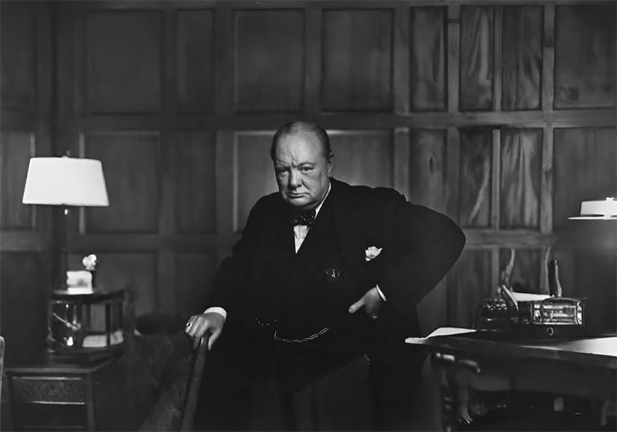 A stern-looking older man in a suit stands with one hand on his hip in a wood-paneled room, next to a desk with papers, a lamp, and a telephone.