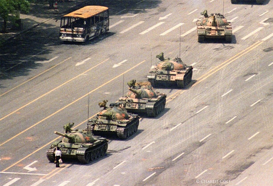A lone person stands in front of a row of four military tanks on a wide city street, with a damaged bus visible in the background. The photo is taken from above.