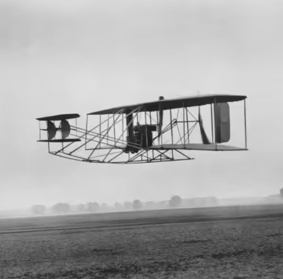 A vintage biplane with a single pilot flies low above an open field, showcasing its exposed framework and twin tail design against a cloudy sky.