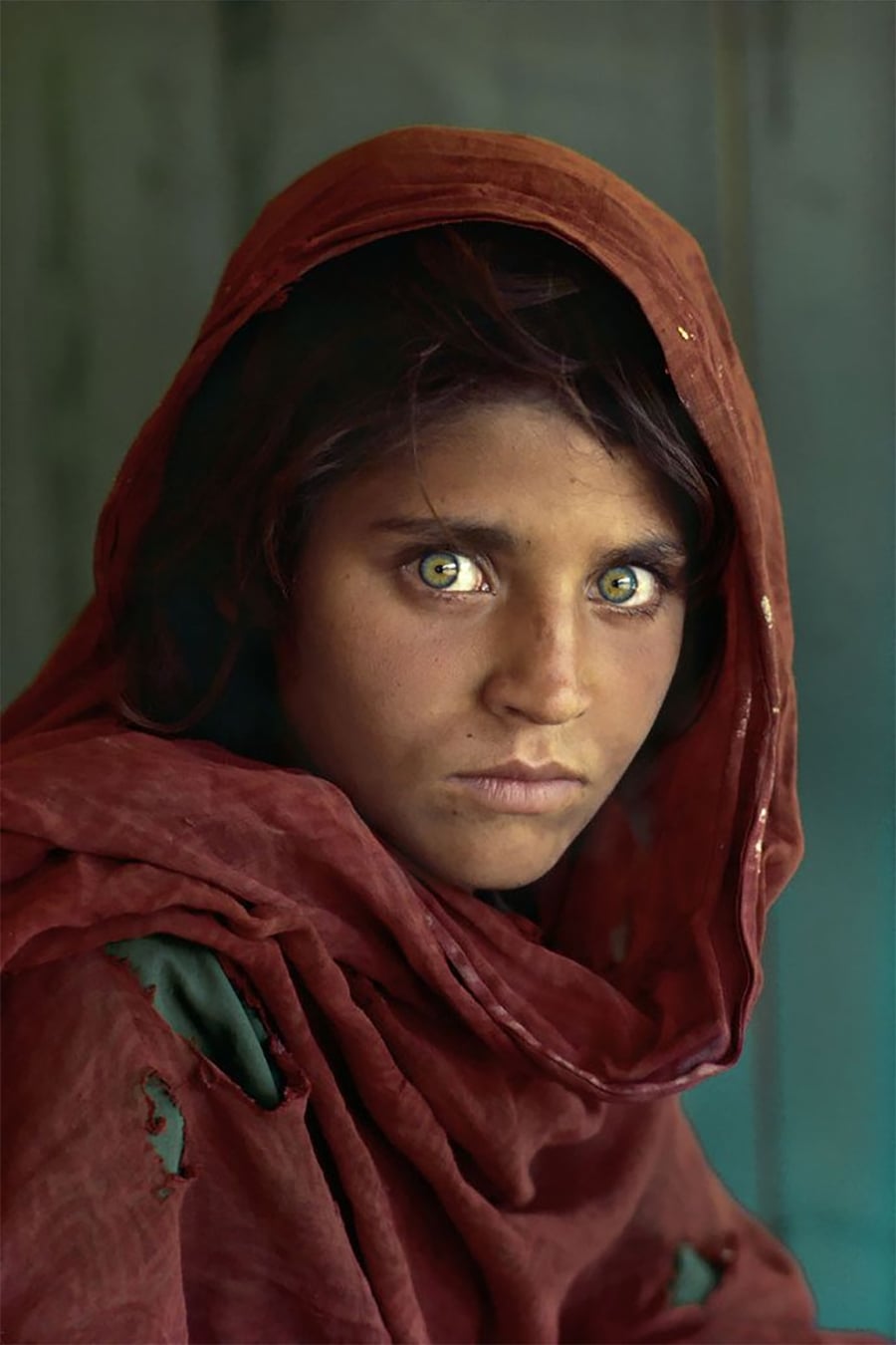 A young girl with striking green eyes, wearing a red headscarf and shawl, looks directly at the camera with an intense expression. The background is a soft green-blue color.