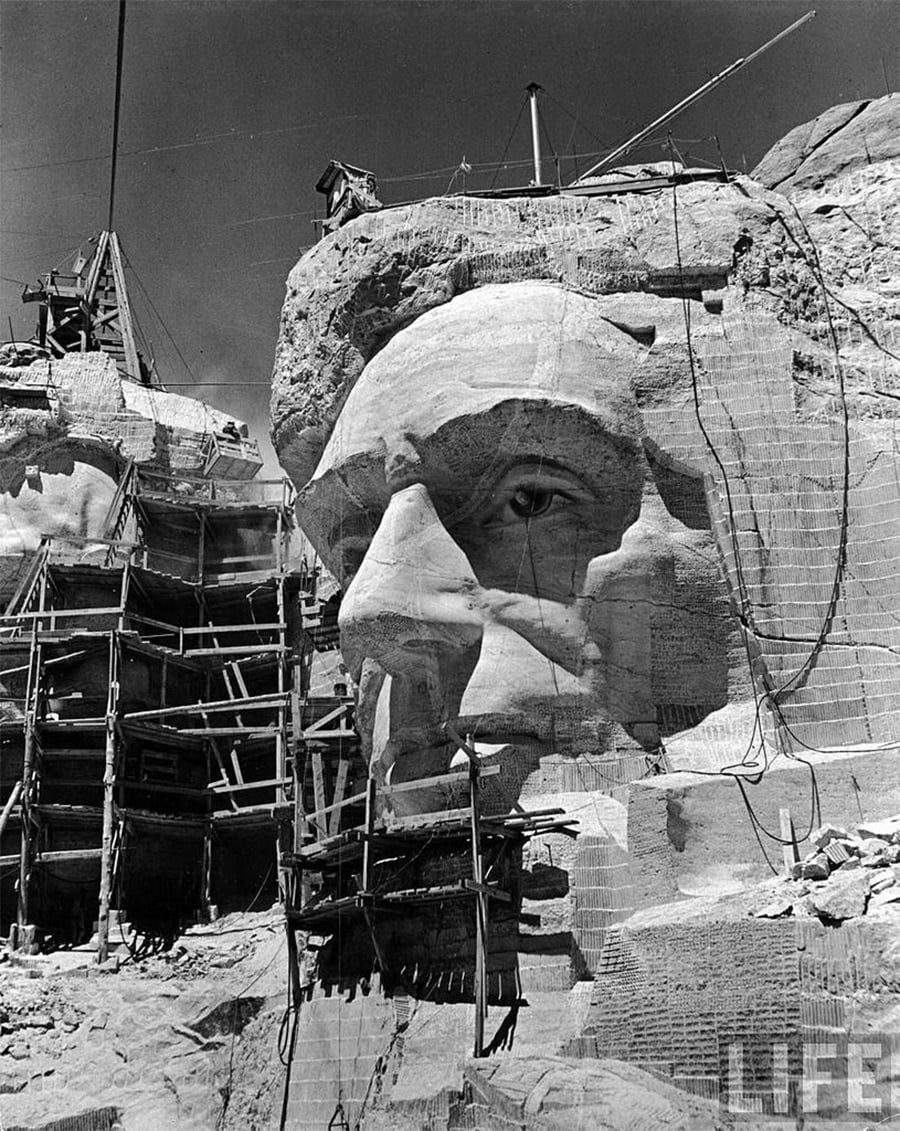 A large stone sculpture of a head is being carved into a mountainside, surrounded by scaffolding and construction equipment under a clear sky.