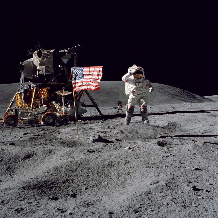 An astronaut stands on the moon near a U.S. flag, lunar module, and lunar rover, with a dark sky and gray, rocky surface in the background.
