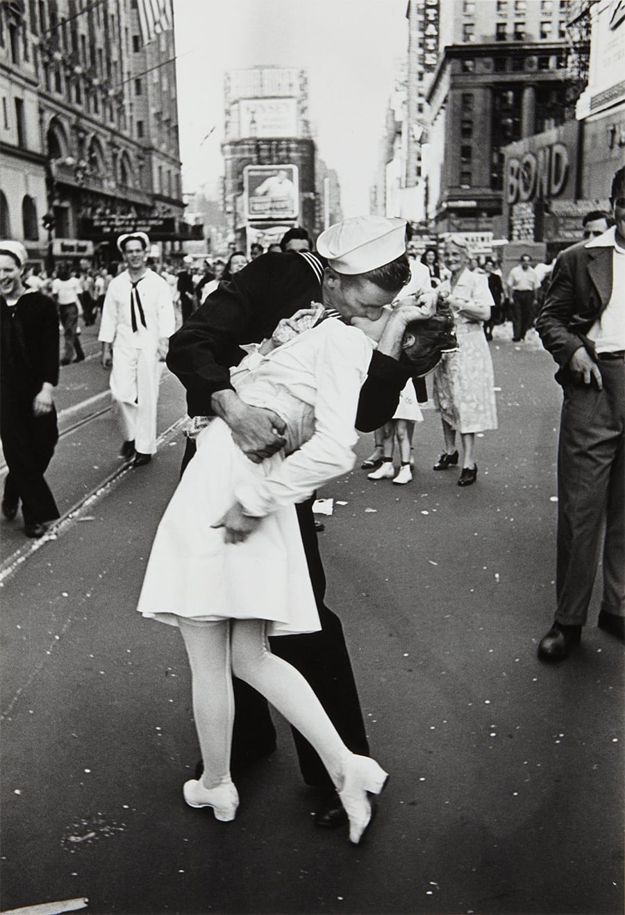 A sailor passionately kisses a nurse in a busy city street, surrounded by people celebrating. The scene is energetic, with buildings and signs visible in the background.