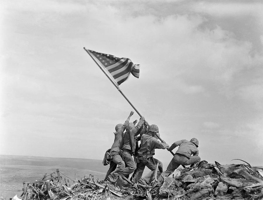 Six soldiers raise an American flag atop a rocky, debris-strewn hill under a cloudy sky during World War II.