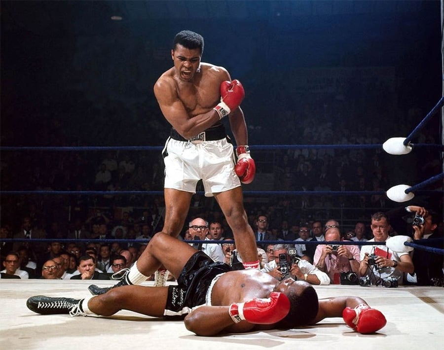 A boxer in white shorts stands over his opponent, who is lying on the mat. The standing boxer gestures with his right arm as spectators and photographers watch from outside the ring.