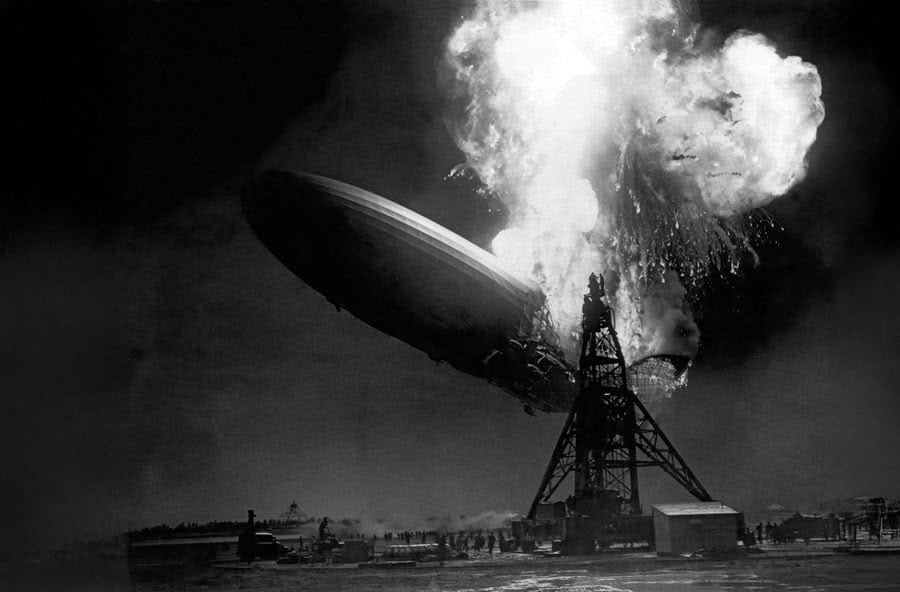 A large airship bursts into flames while moored, with smoke and fire engulfing its rear section. Ground structures and the mooring mast are visible below the disaster.
