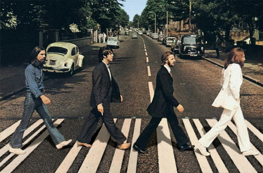 Four men walk in a line across a zebra crossing on a city street, with cars and trees in the background. The first and fourth men wear white, the second and third wear dark suits.