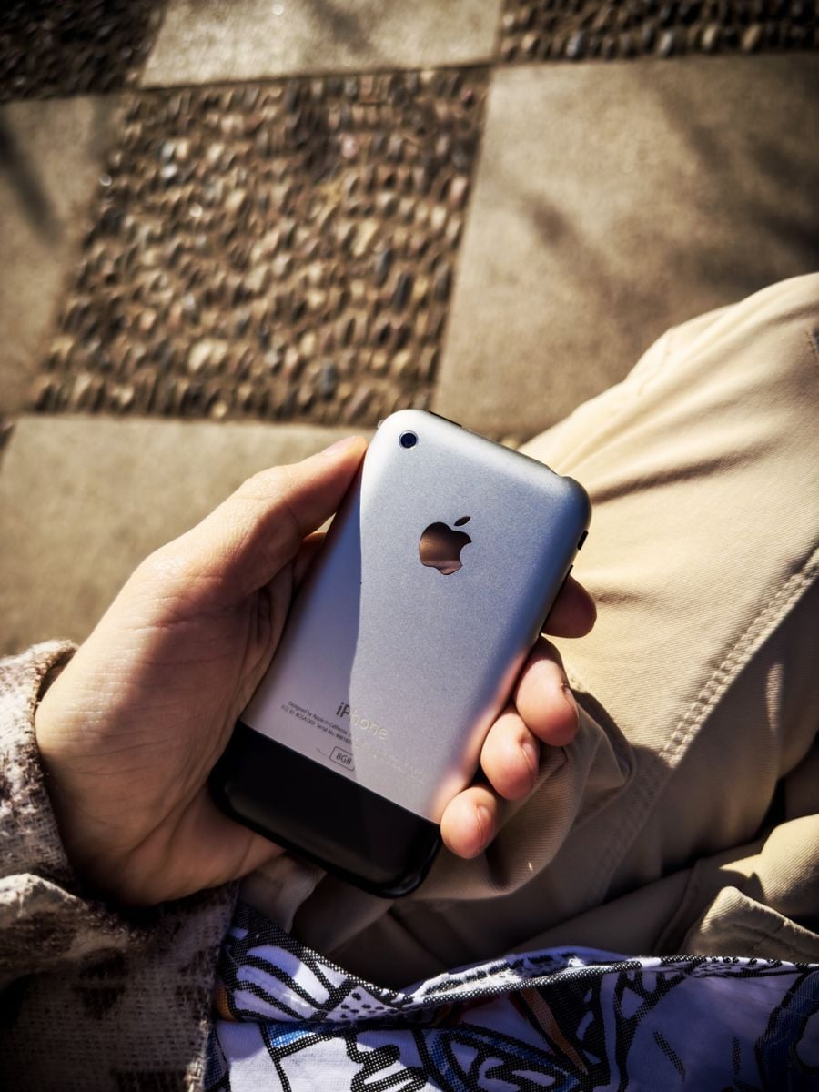 A person holding a classic first-generation iPhone with a silver and black back, sitting outdoors on a sunny tiled surface.