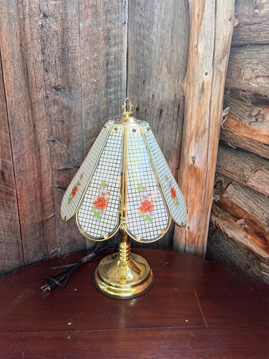 A vintage brass lamp with a stained glass shade featuring pink floral designs sits on a wooden surface against a rustic wooden wall.