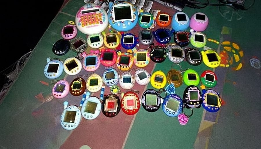 A large collection of various virtual pet devices, including many colorful Tamagotchis, is spread out on a surface. The devices have small screens and different designs.