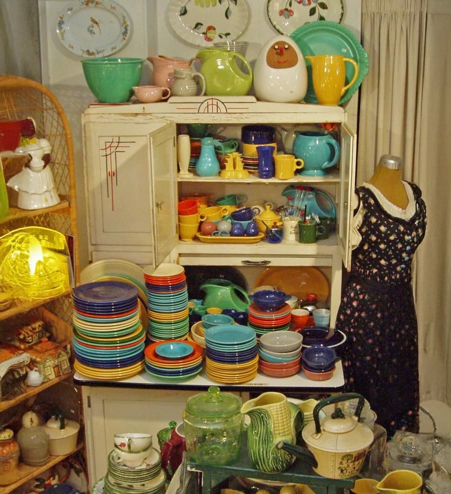 A vintage cabinet filled with colorful dishes, bowls, cups, and teapots. More plates and kitchenware are stacked on tables nearby. A mannequin in a black dress stands to the right. China plates decorate the wall above.