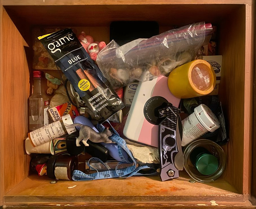 A cluttered wooden drawer containing various items including a tape measure, pill bottles, a yellow cup, a camera, a knife, a vape pack, plastic bags, and other miscellaneous objects.