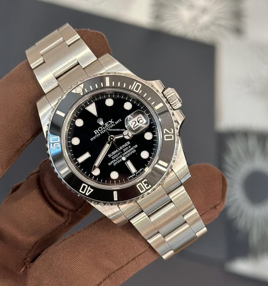 A hand in a brown glove holds a silver Rolex Submariner watch with a black dial and bezel, showing the date at 3 o’clock. The background is blurred with geometric patterns.