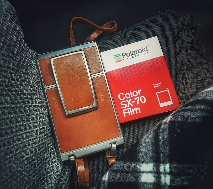 A vintage Polaroid camera with a brown leather case sits next to a red Polaroid Originals Color SX-70 film box on a dark fabric surface.