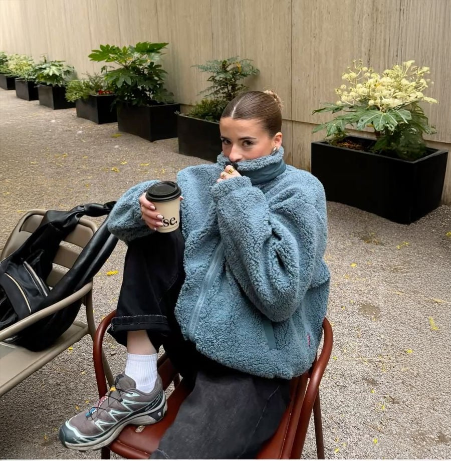 A person in a blue fuzzy jacket sits on a chair holding a coffee cup, with one leg up. They are next to a stroller and black planter boxes with plants, in an outdoor space with a concrete wall.