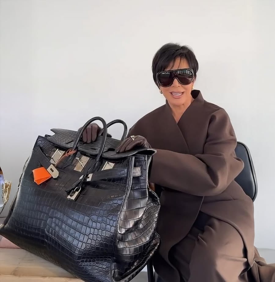 A person in dark sunglasses and a brown coat sits at a table, holding a very large black crocodile-pattern handbag with silver hardware and an orange tag, against a plain white background.