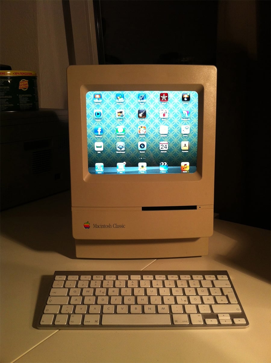 A vintage Macintosh Classic computer displays a modern iPad home screen, with a wireless keyboard in front, on a white desk in a dimly lit room.
