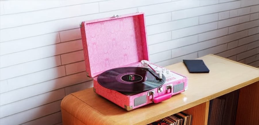 A pink portable record player with a vinyl record sits on a wooden cabinet. A closed notebook or wallet is nearby, and there's a white tiled wall in the background.