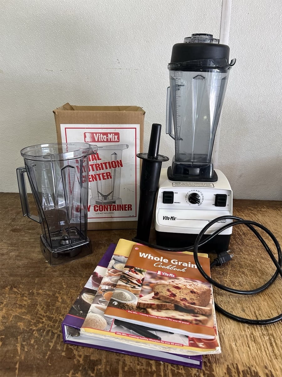 A vintage Vita-Mix blender with an extra pitcher, tamper, instruction manuals, and original box are displayed on a wooden surface.